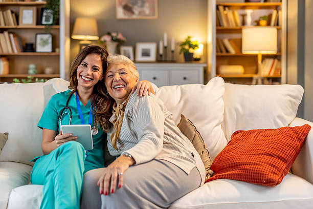 istockphoto-2227949371-612x612 A nurse in scrubs and an elderly woman are smiling while sitting together in a living room, representing health care, companionship, and happiness.